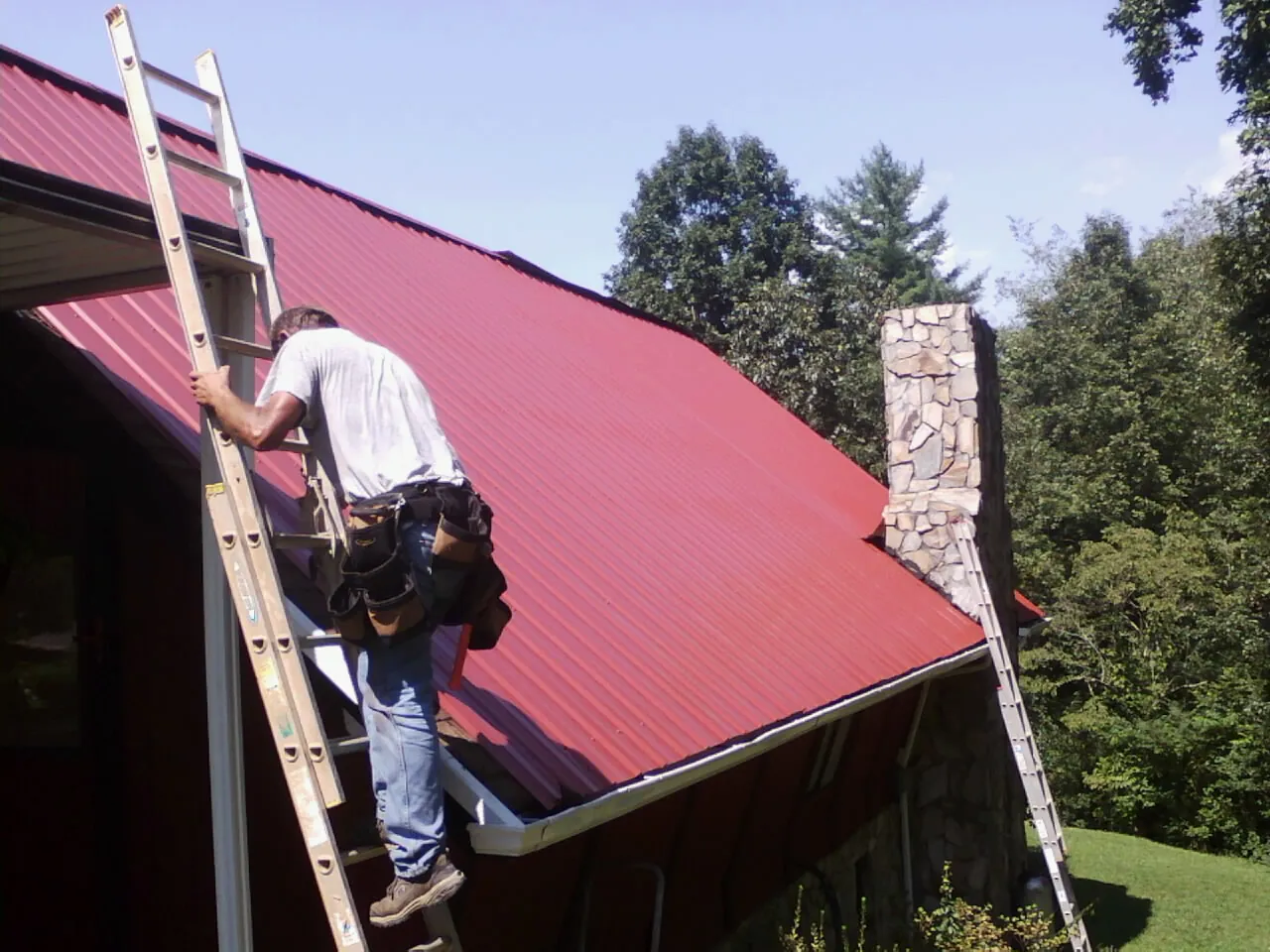 Roofer on a ladder beside a red metal roof and stone chimney — Soffit & Fascia Repair by Roofing Brothers in Lineville
